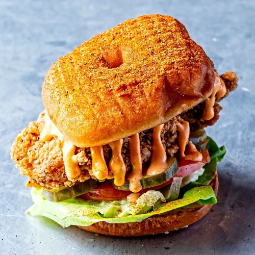 A crispy fried chicken sandwich with a glazed donut bun, topped with lettuce, tomato, pickles, and spicy sauce.