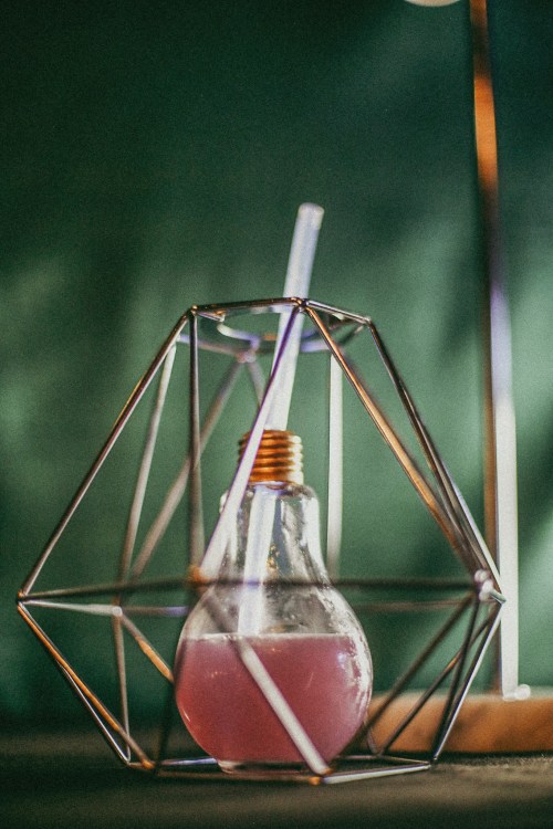 A geometric glass holder containing a pink drink with a straw, set against a green background.
