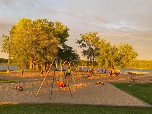 A sandy playground at sunset, with children playing on swings and people enjoying the park by the water.