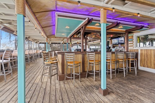 A vibrant bar area with colorful lighting, wooden floors, and high stools, set in a spacious outdoor dining space.