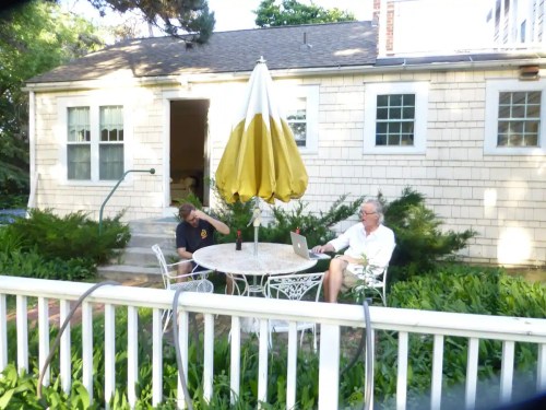 Two people sit at a table under a yellow umbrella in a garden, one using a laptop and the other relaxing.