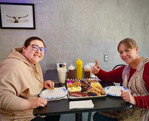 Two women smile at the camera while sitting at a table with a large platter of barbecue and side dishes between them.