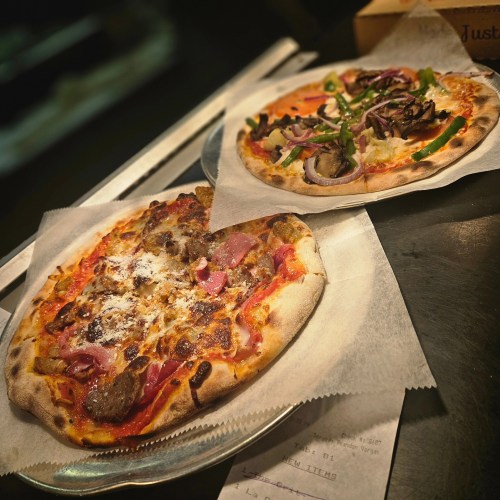Two pizzas on plates: one with meat and cheese, the other topped with vegetables and herbs, on a dark surface.