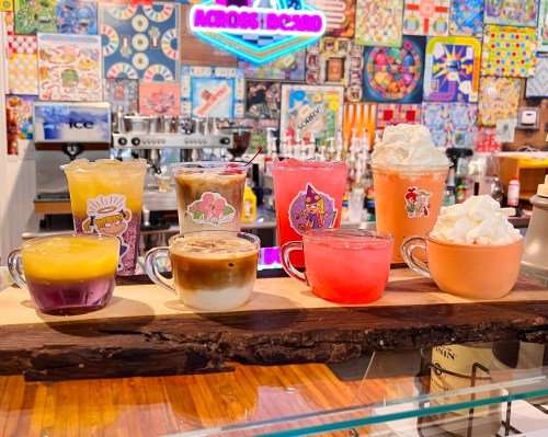 A colorful display of eight drinks in clear cups, each adorned with fun stickers, on a wooden board against a vibrant backdrop.