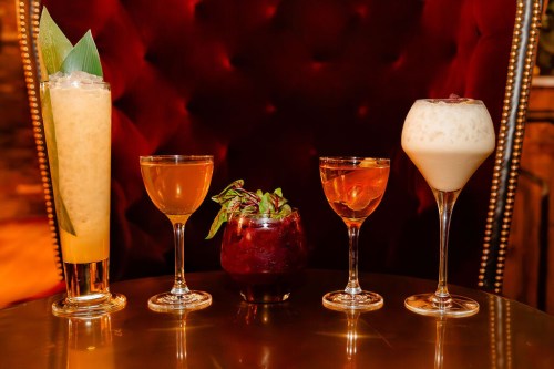 Five colorful cocktails arranged on a table, featuring various glasses and garnishes, set against a plush red background.
