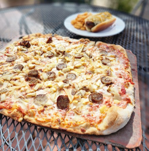 A close-up of a large pizza topped with sausage and pineapple, with a plate of hot dogs in the background.
