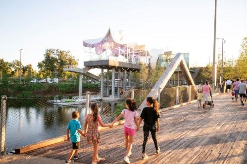 Children walking hand in hand along a wooden path by a river, with a modern building and people in the background.