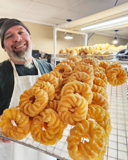 A smiling man in an apron holds a tray of freshly made, golden, spiral-shaped pastries.