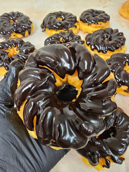 A hand holding a chocolate-glazed donut with a swirl design, surrounded by more donuts on a baking sheet.