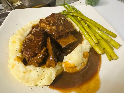 A plate of mashed potatoes topped with beef short ribs in gravy, accompanied by asparagus spears.