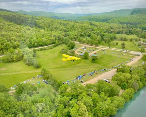 A yellow plane flies over a lush green landscape with a campsite and river below, surrounded by rolling hills.