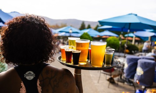A server carries a tray of various beers outdoors, with blue umbrellas and a scenic background.