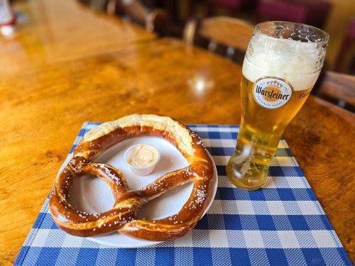 A large pretzel with salt on a plate, accompanied by a small bowl of dip, next to a glass of beer on a checkered tablecloth.