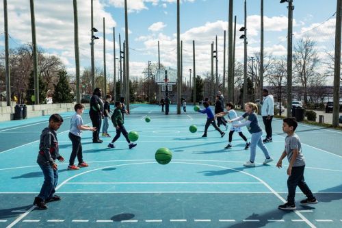 Children and adults play basketball on a blue court, with several green balls in motion under a partly cloudy sky.