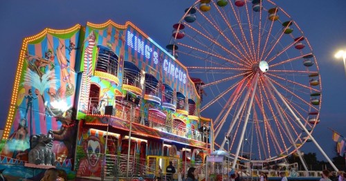 A colorful carnival scene featuring a circus-themed ride and a large Ferris wheel against a twilight sky.