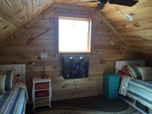 Cozy attic bedroom with two beds, wooden walls, a window, and colorful decor including a lamp and storage unit.