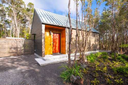 A modern cabin with a metal roof and orange door, surrounded by trees and landscaped gravel.