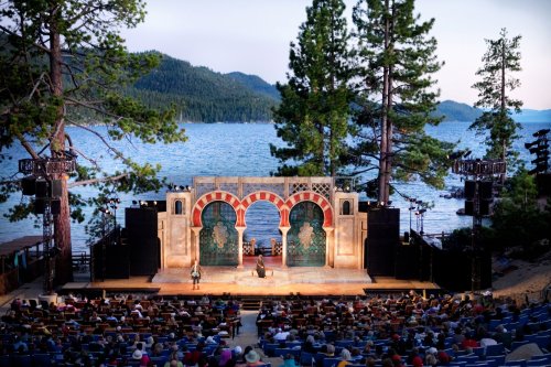 An outdoor theater by a lake, with a scenic backdrop of trees and mountains, hosting a performance for a large audience.
