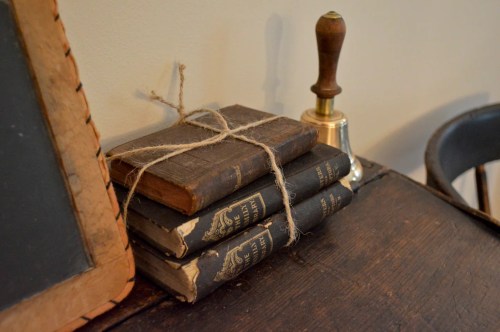 A stack of old books tied with twine next to a bell and a chalkboard on a wooden table.