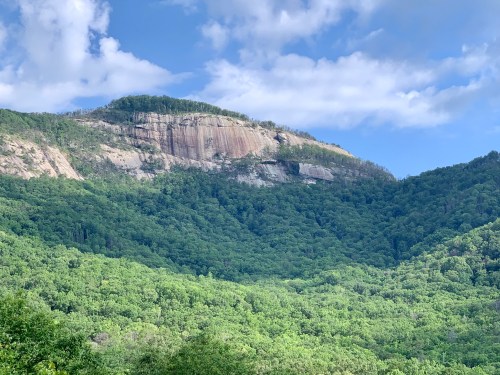 A rocky mountain rises above a lush green forest under a partly cloudy blue sky.
