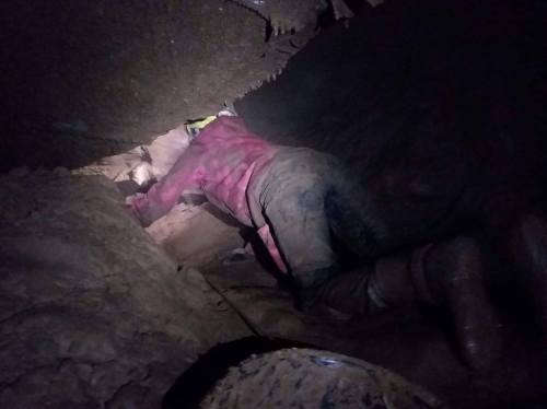 A person in muddy clothing crawls through a narrow, dark cave passage, using a headlamp for light.