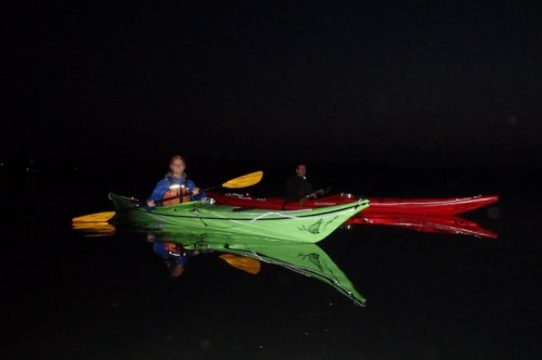 Two kayakers in green and red kayaks on calm water at night, with reflections visible in the water.