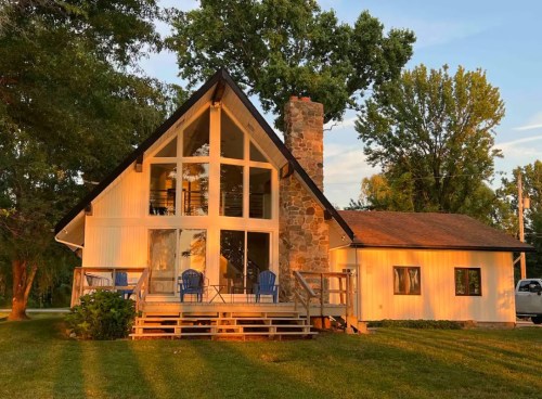 A charming A-frame house with a stone chimney, large windows, and a wooden deck surrounded by trees at sunset.