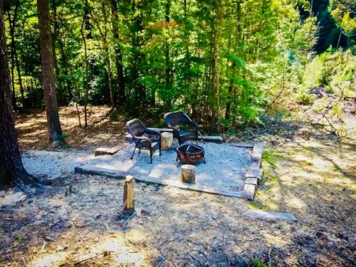 A cozy fire pit area in a wooded setting, featuring two chairs and a stone circle surrounded by trees.
