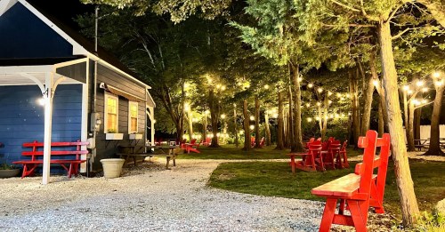 A cozy outdoor space at night with red chairs, string lights, and a blue building surrounded by trees.