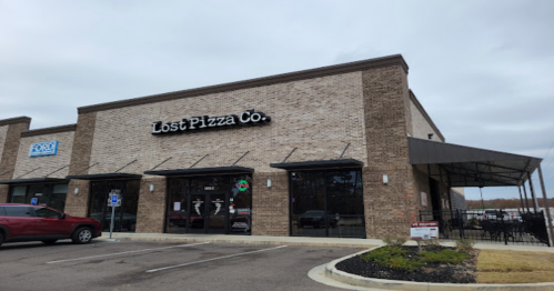Exterior of Lost Pizza Co. restaurant, featuring a brick facade and outdoor seating area, under a cloudy sky.