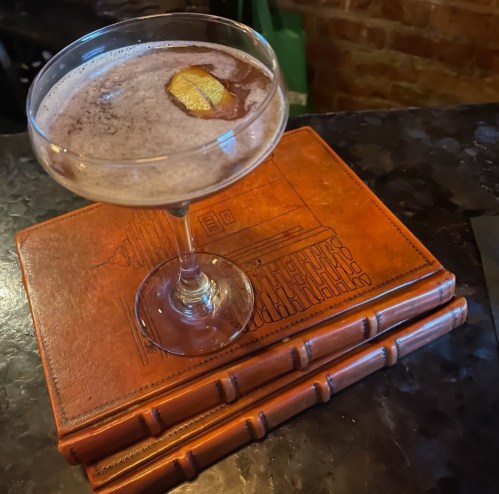 A cocktail in a coupe glass sits atop stacked leather-bound books on a dark countertop.