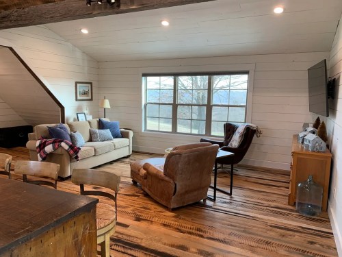 Cozy living room with a sofa, armchair, and large window showcasing a scenic view. Warm wood floors and rustic decor.