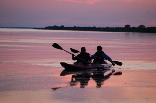 Two people kayaking on a calm lake at sunset, with vibrant pink and purple reflections on the water.