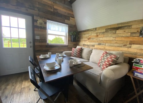 Cozy interior of a small room with a sofa, dining table set for four, and wooden walls. Natural light from the door.