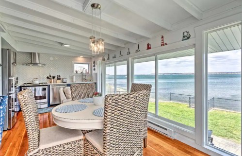 Bright dining area with a round table and woven chairs, overlooking a scenic waterfront through large windows.