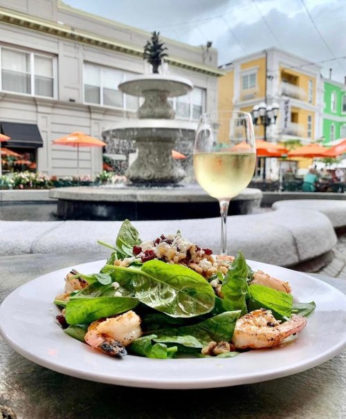 A fresh spinach salad with shrimp and cranberries, accompanied by a glass of white wine, set near a fountain.
