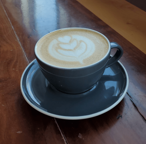 A gray cup of latte art with a heart design, sitting on a matching saucer on a wooden table.