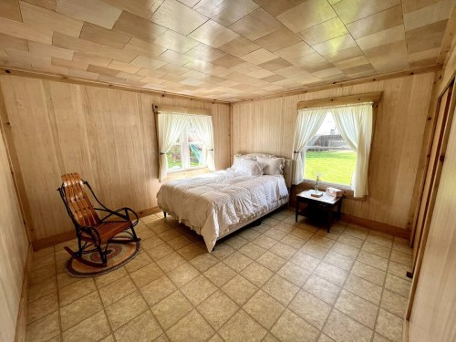 Cozy bedroom with a bed, rocking chair, and two windows letting in natural light. Light wood paneling and tiled floor.