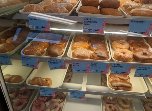 A display case filled with various donuts, including glazed, filled, and specialty flavors, with price tags visible.
