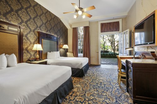A hotel room with two beds, elegant decor, a desk, and a view of a garden through open doors.