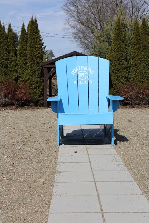 A large blue Adirondack chair sits on a stone path, surrounded by greenery and trees in a sunny outdoor setting.