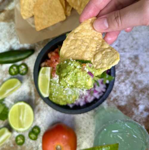 A hand holding a tortilla chip dipped in guacamole, surrounded by lime wedges, jalapeños, and a bowl of salsa.