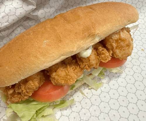 A sandwich with a soft bun filled with fried chicken, lettuce, and tomato, served on a patterned paper.