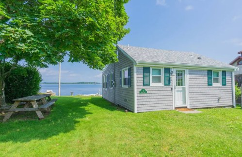 A cozy cottage by the water, surrounded by green grass and trees, with a picnic table nearby.