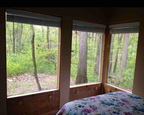 Cozy room with three large windows overlooking a lush green forest. A colorful quilt is on the bed.