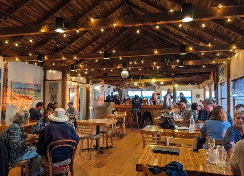 A cozy restaurant interior with wooden beams, string lights, and diners enjoying meals at wooden tables.