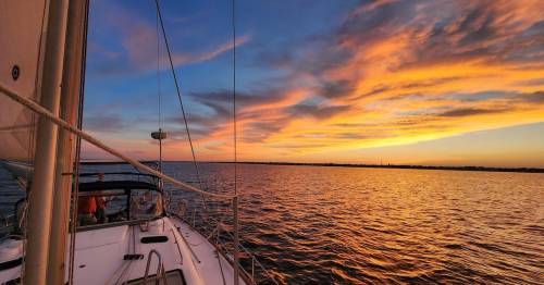 A sailboat on calm waters at sunset, with vibrant orange and purple clouds reflecting on the water's surface.