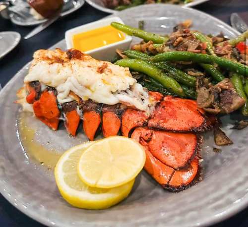 A plate featuring a lobster tail with lemon slices, green beans, and a small dish of sauce.