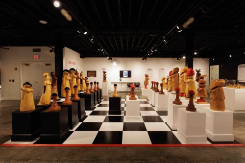 An art exhibition featuring wooden sculptures displayed on pedestals in a checkerboard pattern.