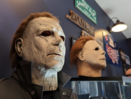 Two realistic Michael Myers masks displayed on a shelf against a dark blue wall, with signs and decor in the background.
